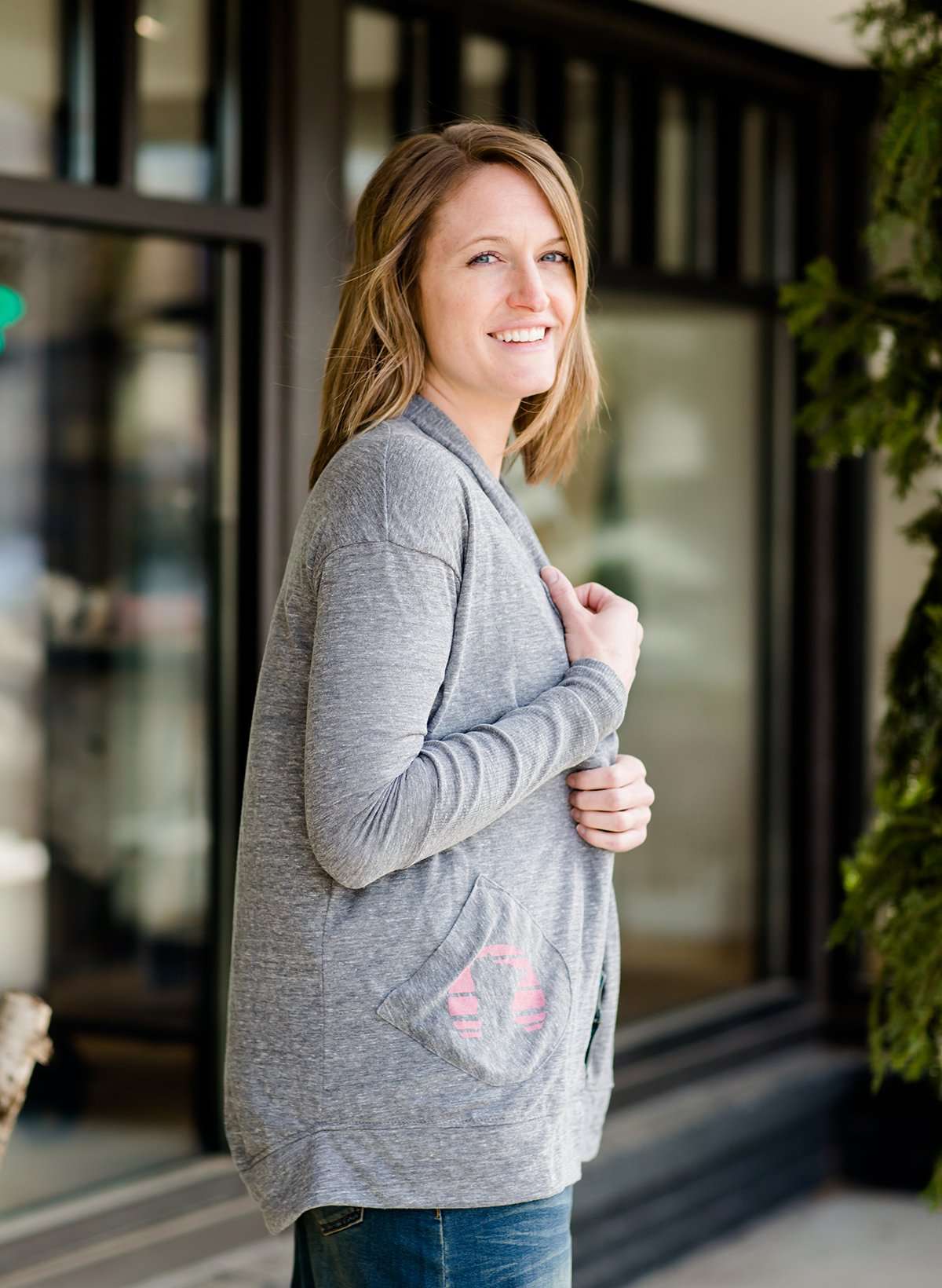 Woman standing in front of Inherit Clothing Company wearing a Minnesota screen printed, gray, open cardigan. The Minnesota screen print is pink.