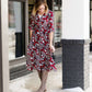 Woman wearing a midi length modest dress. This dress is a holiday red with poppy flowers and white flowers on it. It has a v-neck and ruffle 3/4 sleeves. This dress is paired with tall suede riding boots.