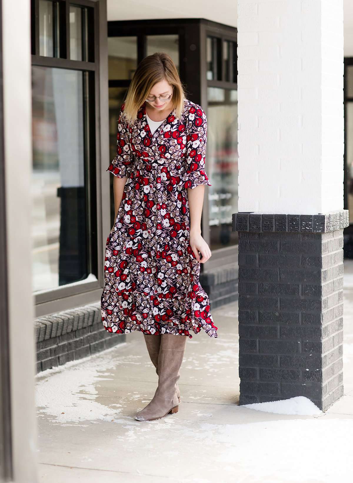Woman wearing a midi length modest dress. This dress is a holiday red with poppy flowers and white flowers on it. It has a v-neck and ruffle 3/4 sleeves. This dress is paired with tall suede riding boots.