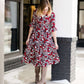 Woman wearing a midi length modest dress. This dress is a holiday red with poppy flowers and white flowers on it. It has a v-neck and ruffle 3/4 sleeves. This dress is paired with tall suede riding boots.