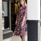 Woman wearing a midi length modest dress. This dress is a holiday red with poppy flowers and white flowers on it. It has a v-neck and ruffle 3/4 sleeves. This dress is paired with tall suede riding boots.