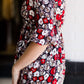 Woman wearing a midi length modest dress. This dress is a holiday red with poppy flowers and white flowers on it. It has a v-neck and ruffle 3/4 sleeves. This dress is paired with tall suede riding boots.