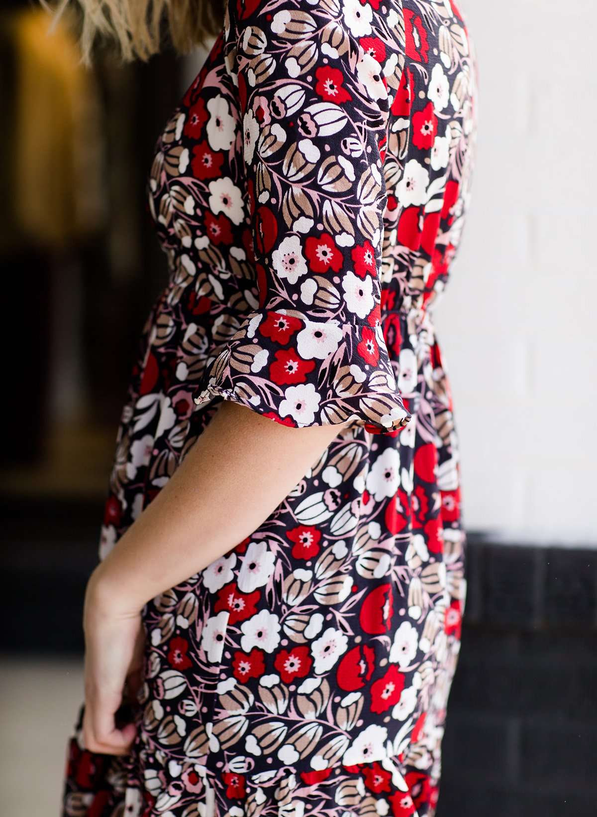 Woman wearing a midi length modest dress. This dress is a holiday red with poppy flowers and white flowers on it. It has a v-neck and ruffle 3/4 sleeves. This dress is paired with tall suede riding boots.