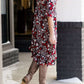 Woman wearing a midi length modest dress. This dress is a holiday red with poppy flowers and white flowers on it. It has a v-neck and ruffle 3/4 sleeves. This dress is paired with tall suede riding boots.