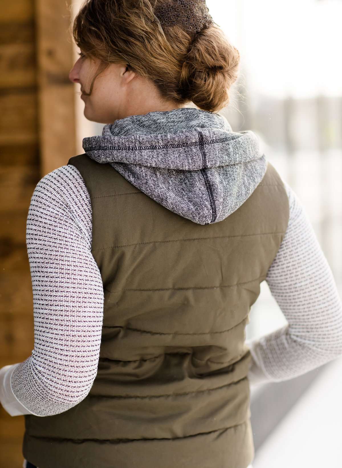Woman wearing an anorak quilted vest with a zipper and hood. This vest comes in olive or black with gray accents.